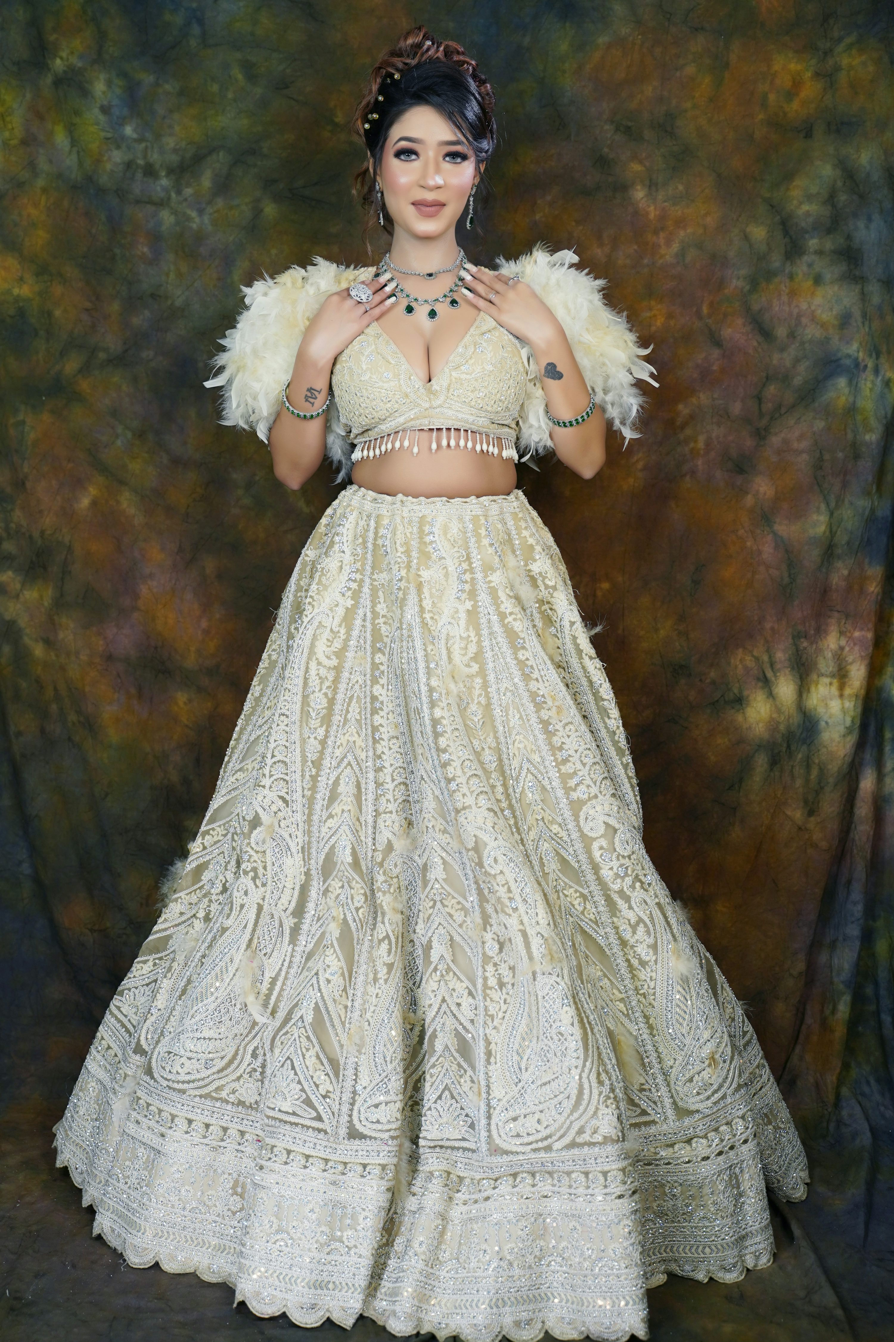 a fulllength studio portrait of a young woman posing in a beautiful, elaborate offwhite lehenga choli. the dress features intricate embroidery and lace detailing throughout. she's accessorized with a matching feather shawl, a necklace with emerald gemstones, and various rings and bangles. her makeup is sophisticated and she wears her hair in a styled updo. the backdrop is a textured, dark brown and gold color. the overall image conveys elegance, sophistication, and bridal style.