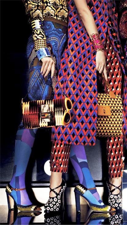 he image shows two women's legs, dressed in vibrant and patterned clothing and accessories. the women are wearing colorful tights with geometric designs, highheeled platform shoes with floral prints, and carrying patterned handbags. their clothing features a mix of geometric and floral patterns in bold, bright colors. the overall style is reminiscent of retro or vintage fashion trends from the 1960s or 70s, with a psychedelic feel. the image is a fashion photograph, showcasing the clothing and accessories.