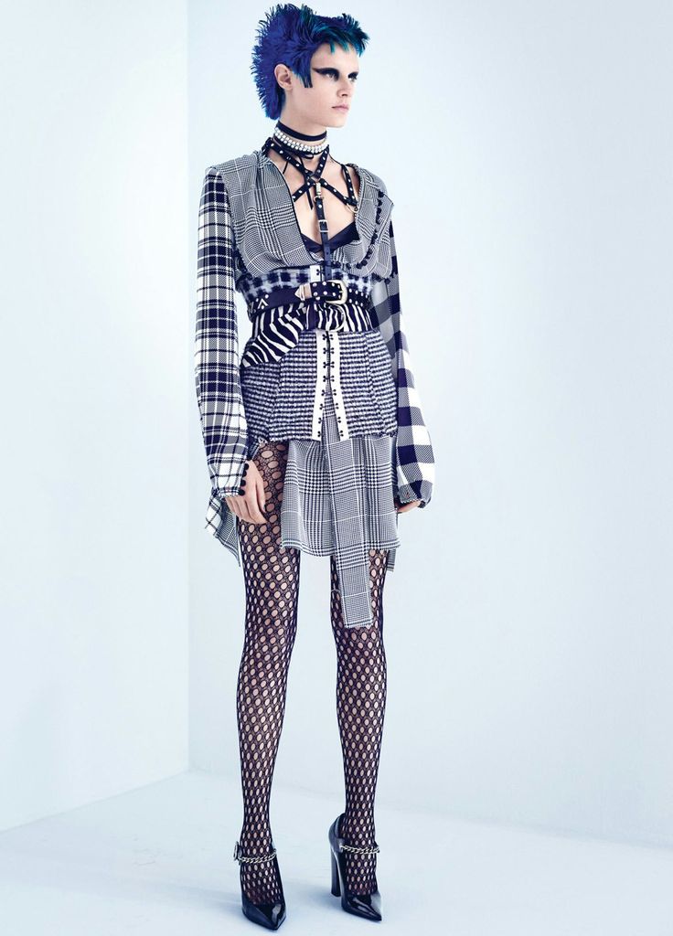 a woman with blue spiked hair is wearing a punkstyle fashion outfit. she is wearing a plaid dress with a zebraprint belt, fishnet stockings, and high heels with chains. she also has a choker and a harness. the background is a bright white corner.