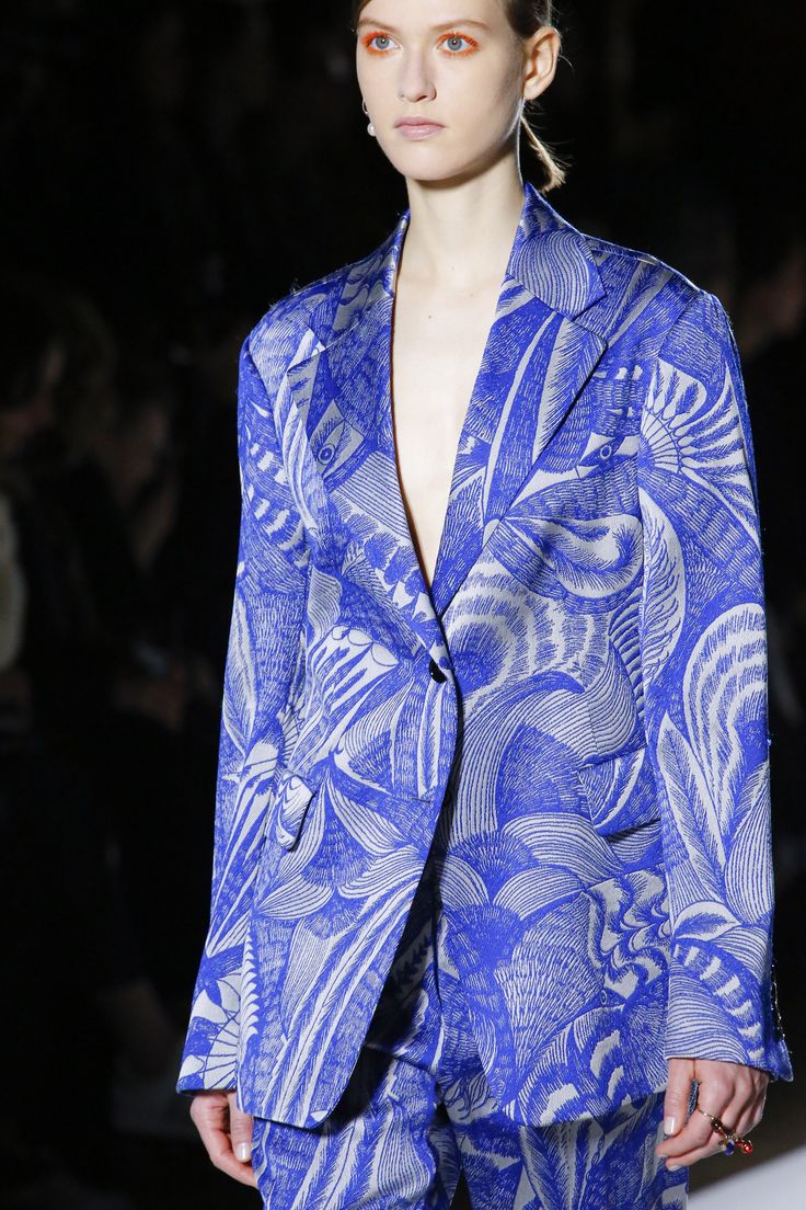 loseup view of a female fashion model showcasing a vibrant blue suit. the suit features a striking, detailed print that resembles stylized birds or floral motifs in shades of blue and white on a  blue background. the model has her hair pulled back, and orangetoned makeup is noticeable. she appears to be wearing simple earrings and a ring. the background is blurred and out of focus, suggesting a fashion show or runway setting. the image focuses on the texture and design of the suit.
