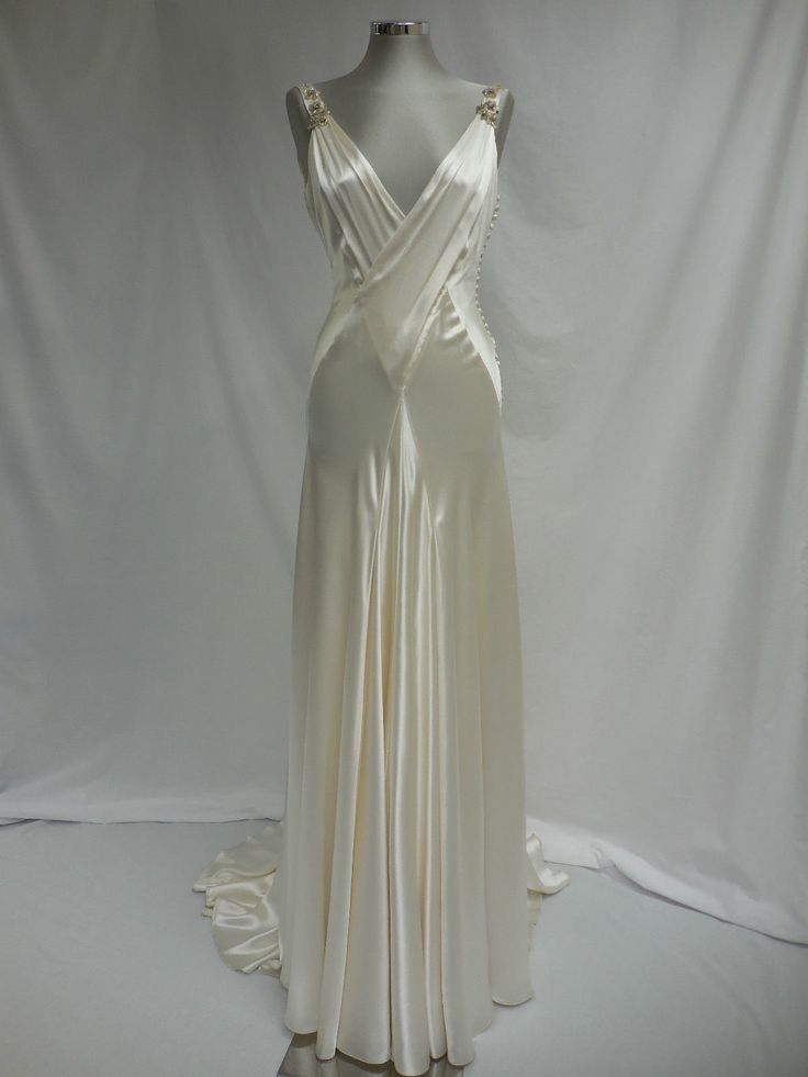 a full shot of a creamcolored satin wedding dress on a mannequin. the dress has a vneck, draped bodice, and a long, flowing skirt with a small train. the dress appears to be a vintage style.