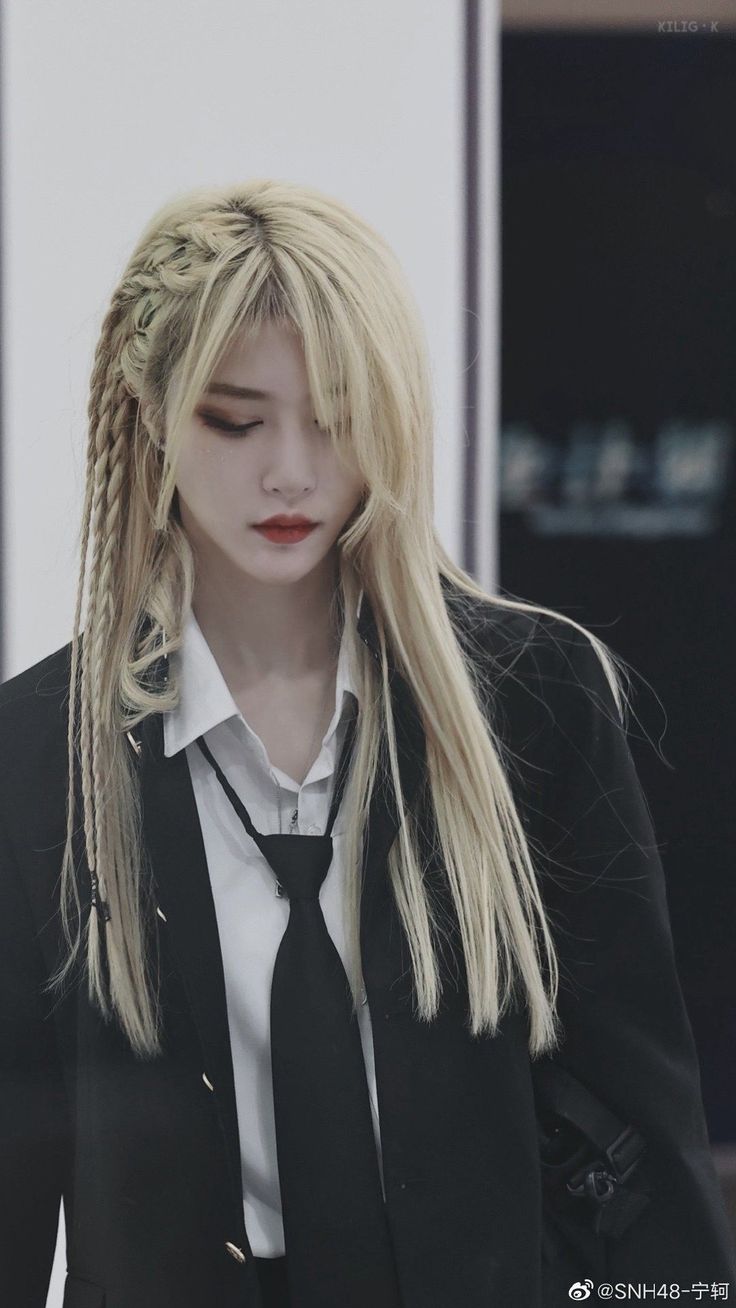 a young woman with blonde hair styled in braids is the subject of this image. she's wearing a black blazer, white collared shirt, and black tie. her makeup is minimal with a bold red lip. the background suggests an indoor environment with soft lighting.