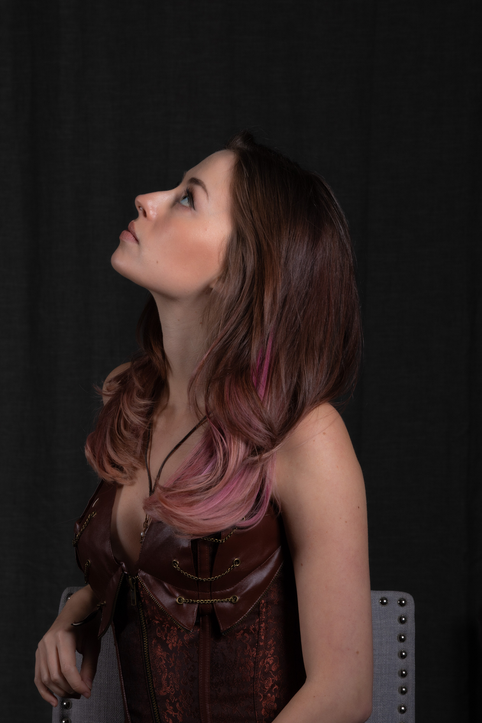 a young woman with pink highlights in her hair poses in steampunk attire.  she is wearing a brown leather and brocade corset and is turned to the side, looking upward. the background is solid black.