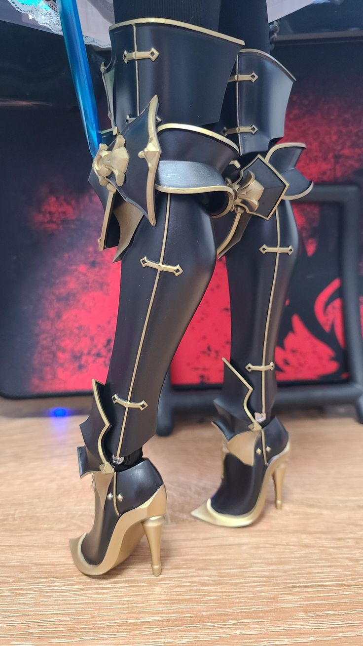 a pair of black and gold thighhigh boots with intricate armor details, worn by a figurine or action figure. the boots have a high heel and a sleek, stylish design. the figure appears to be wearing other armor pieces as well, suggesting a fantasy or cosplay theme. the boots are the main focus of the image, showcasing their intricate details and unique design.