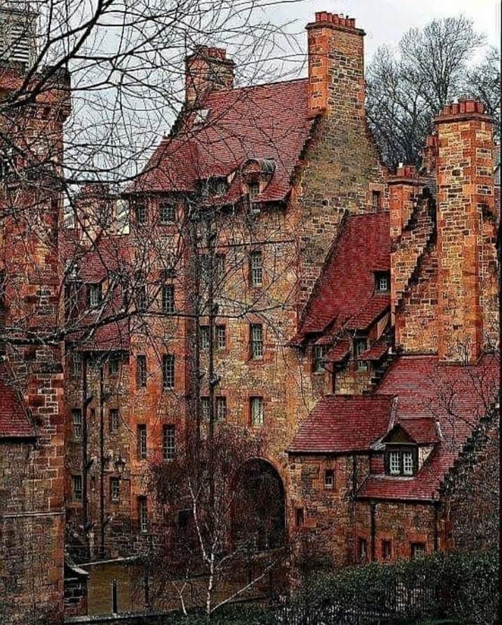 the image shows an old brick building, likely in europe. the building is made of red and brown bricks and has a lot of windows. there is an archway in the center of the building. there are trees in front of the building, and the branches of the trees are bare. the sky is visible in the background. the colors in the image are mostly reds, browns, and greens. the lighting is natural and the perspective is eye level.