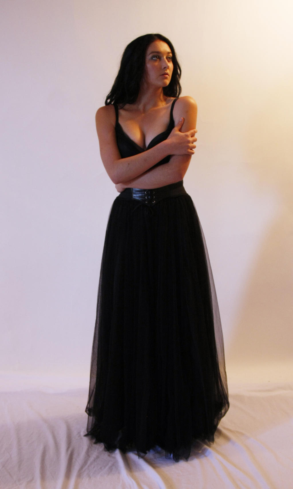 a fulllength shot of a young woman with long dark hair standing against a white background. she is wearing a black tulle maxi skirt with a black corset belt, and a black bra top. her arms are crossed over her chest, and she is looking away from the camera. the lighting is soft and even.
