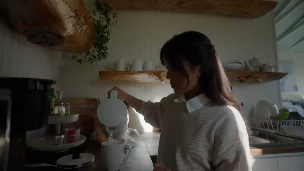 a woman in a white sweater is standing in a brightly lit kitchen with white walls and wooden accents. she is pouring a hot drink, likely coffee or tea, from a white electric kettle into a white mug. the image evokes a sense of calm and peacefulness, hinting at a cozy morning routine.