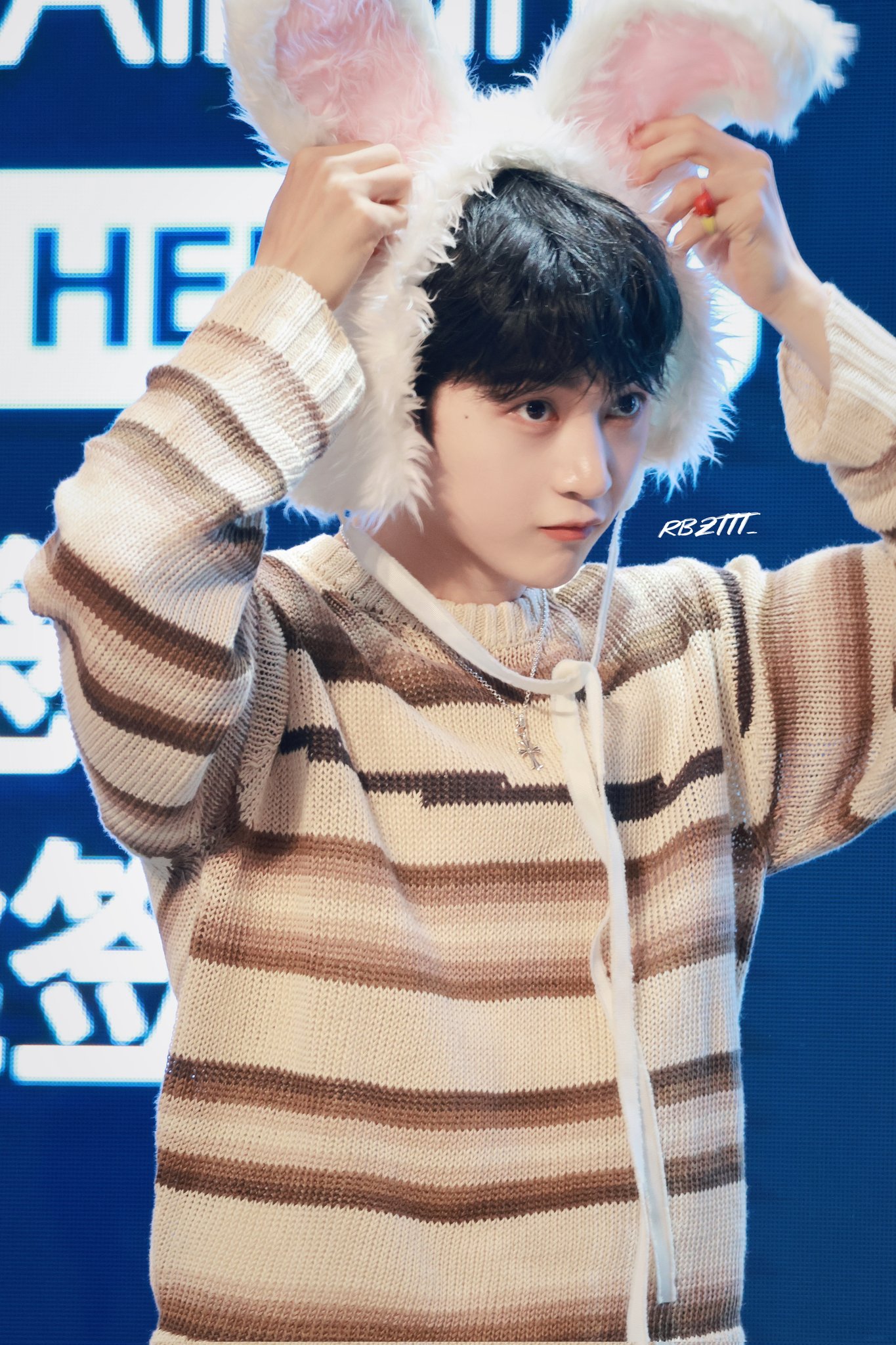 the image shows a young asian man indoors, wearing a striped, knit sweater and white, fluffy bunny ears on his head. he is looking slightly down and to the right, with a subtle smile on his face. the background features blurred blue and white signage, and the lighting appears to be artificial indoor lighting.