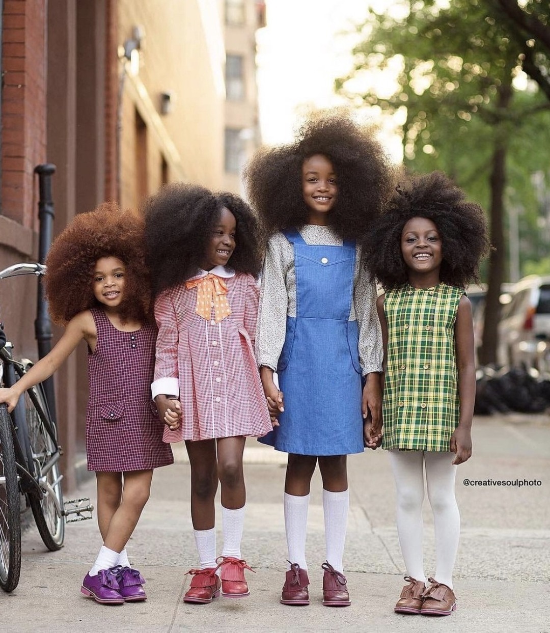 four girls are posing in a street, smiling and looking at the camera. they are all wearing beautiful dresses, the first girl is wearing a purple checked dress, second girl is wearing a pink dress, the third girl is wearing a denim dress, and the fourth girl is wearing a green plaid dress. they all have gorgeous afro hair.
