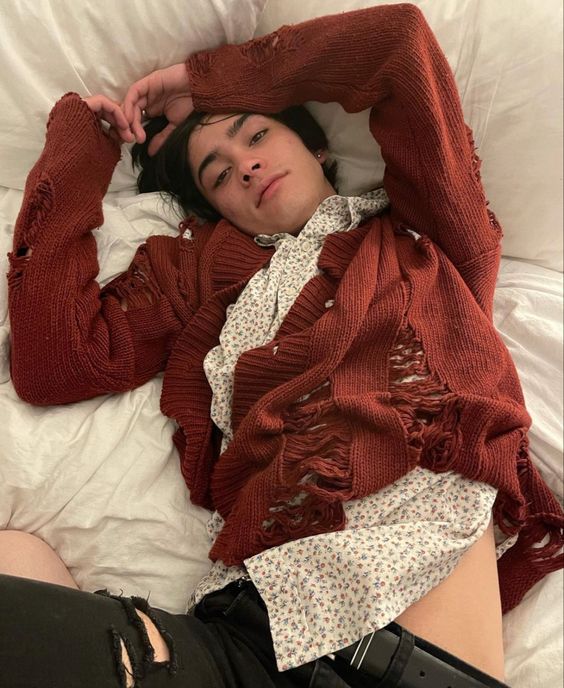 young man is sprawled out on a bed, looking directly at the camera. he is wearing a distressed, oversized orangebrown cardigan over a white shirt with a small floral print, ripped black jeans, and a black belt. the room is brightly lit with natural light, suggesting daytime.  the photo is taken from a straighton perspective.