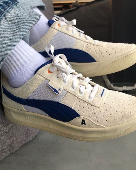the image shows a pair of puma sneakers being worn. the sneakers have a suede upper in an offwhite or cream color, with blue accents and white laces. the wearer is also wearing white socks and blue jeans.