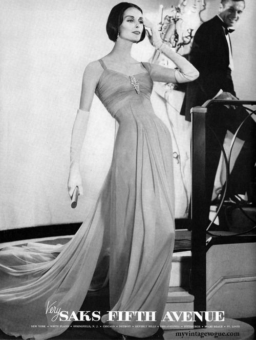 a black and white advertisement for saks fifth avenue featuring a woman in a glamorous evening gown with a flowing train. she stands in front of a man who is walking up a staircase. the image evokes a sense of elegance and sophistication.