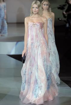 two fashion models are walking on a runway, one in a pink and white gown, the other in a white gown. both models are wearing their hair down and have natural makeup. the runway is made of a light grey material and the background is a blurry white. the models are posed in a way that highlights the fabric of their gowns. the lighting is soft and even, creating a glamorous atmosphere. the photo is taken from a low angle, which gives the models a sense of grandeur.