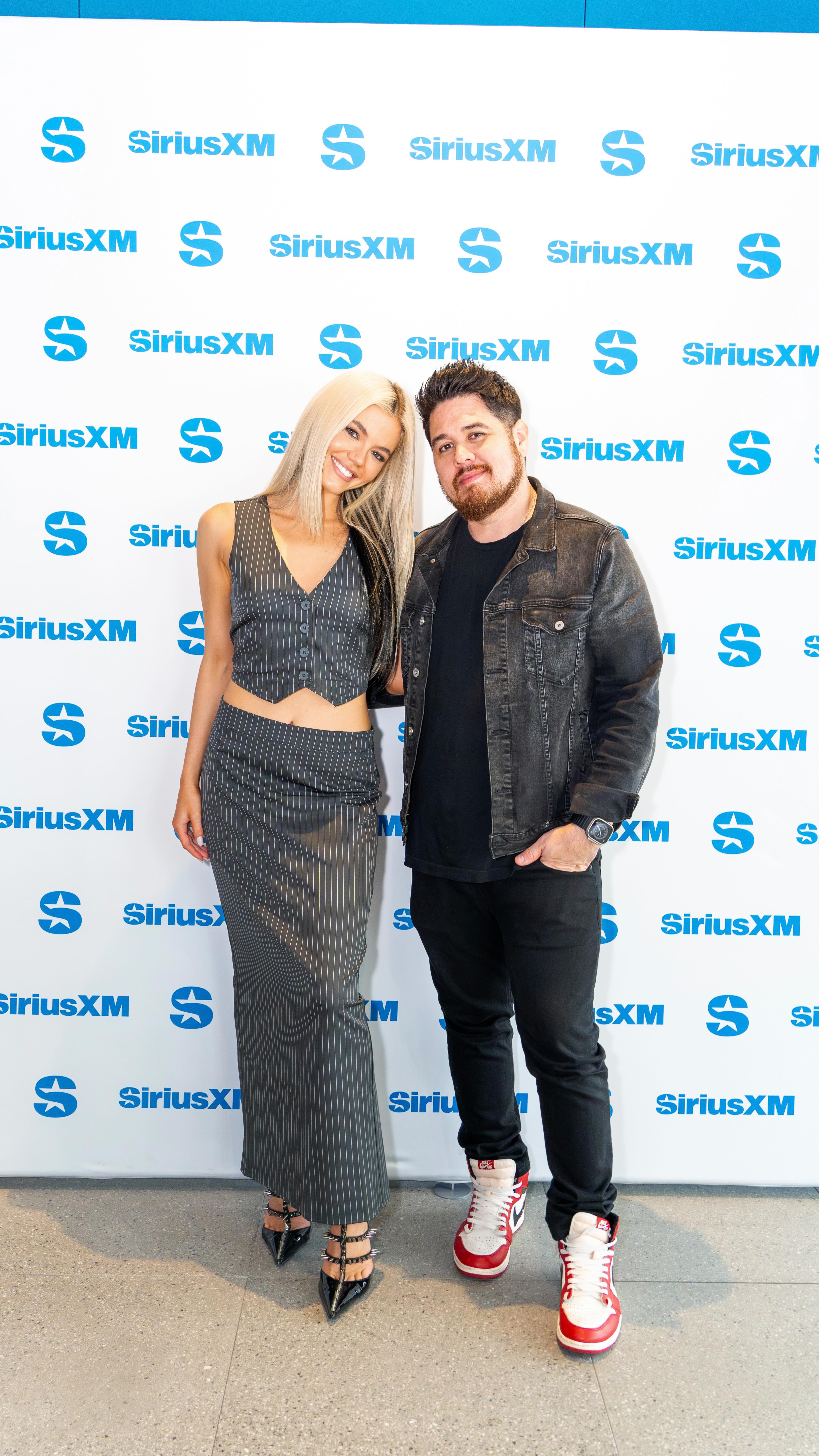 two people, a woman and a man, are standing in front of a backdrop with the siriusxm logo repeated across it. the woman has blonde hair and is wearing a matching pinstripe vest and long skirt, paired with black pointed heels. the man is wearing a black tshirt, black jeans, a dark denim jacket, and red and white sneakers.