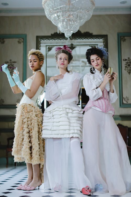 hree women in elaborate 18thcenturystyle dresses stand in a grand, antique interior. the dresses are in pastel shades of white, beige, and pink, and feature ruffled skirts and intricate detailing. the women wear wigs or elaborate hairstyles and are holding accessories such as perfume bottles and a powder puff.  the setting appears to be a luxurious room with ornate mirrors and a large crystal chandelier. the overall style is elegant and reminiscent of the rococo period.