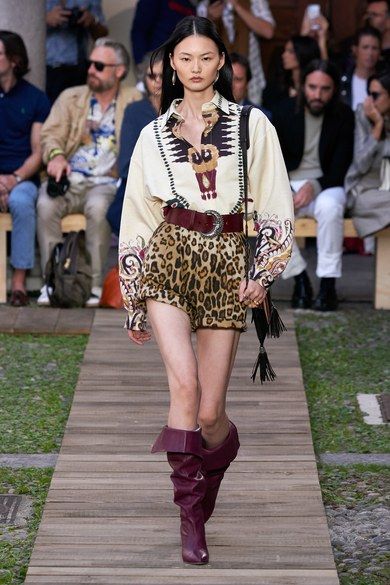 a fashion model walks down a runway wearing a stylish outfit.  the outfit consists of a creamcolored, longsleeved shirt with native americaninspired embroidery, leopard print shorts, a brown leather belt with a large buckle, and tall, slouchy burgundy boots. the model also carries a fringed handbag. the background features other people seated as an audience, suggesting a fashion show setting. the overall style is a blend of bohemian, western, and modern elements. the runway is made of wood.