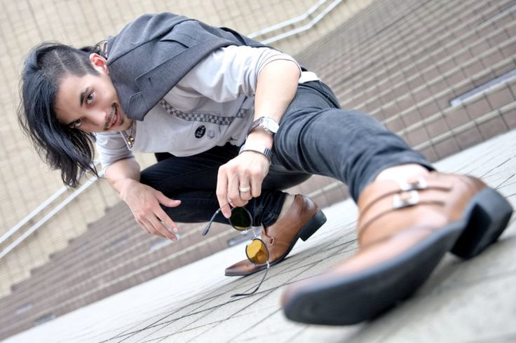 a man in a stylish, casual outfit, with his left leg bent and right leg extended. the image is taken from a low angle, showcasing the model's footwear and the overall style of the outfit. he is holding a pair of sunglasses in his hand,  and the shot gives a cool, trendy vibe.