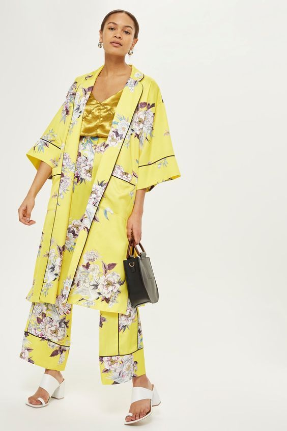 a fullbody shot of a model wearing a yellow floral kimono and matching wideleg pants.  the kimono and pants are made of a satinlike material and feature a largescale floral print. she is also wearing a gold camisole underneath the kimono, black handbag, and white heeled sandals. the background is plain white. the overall style is summery and elegant.