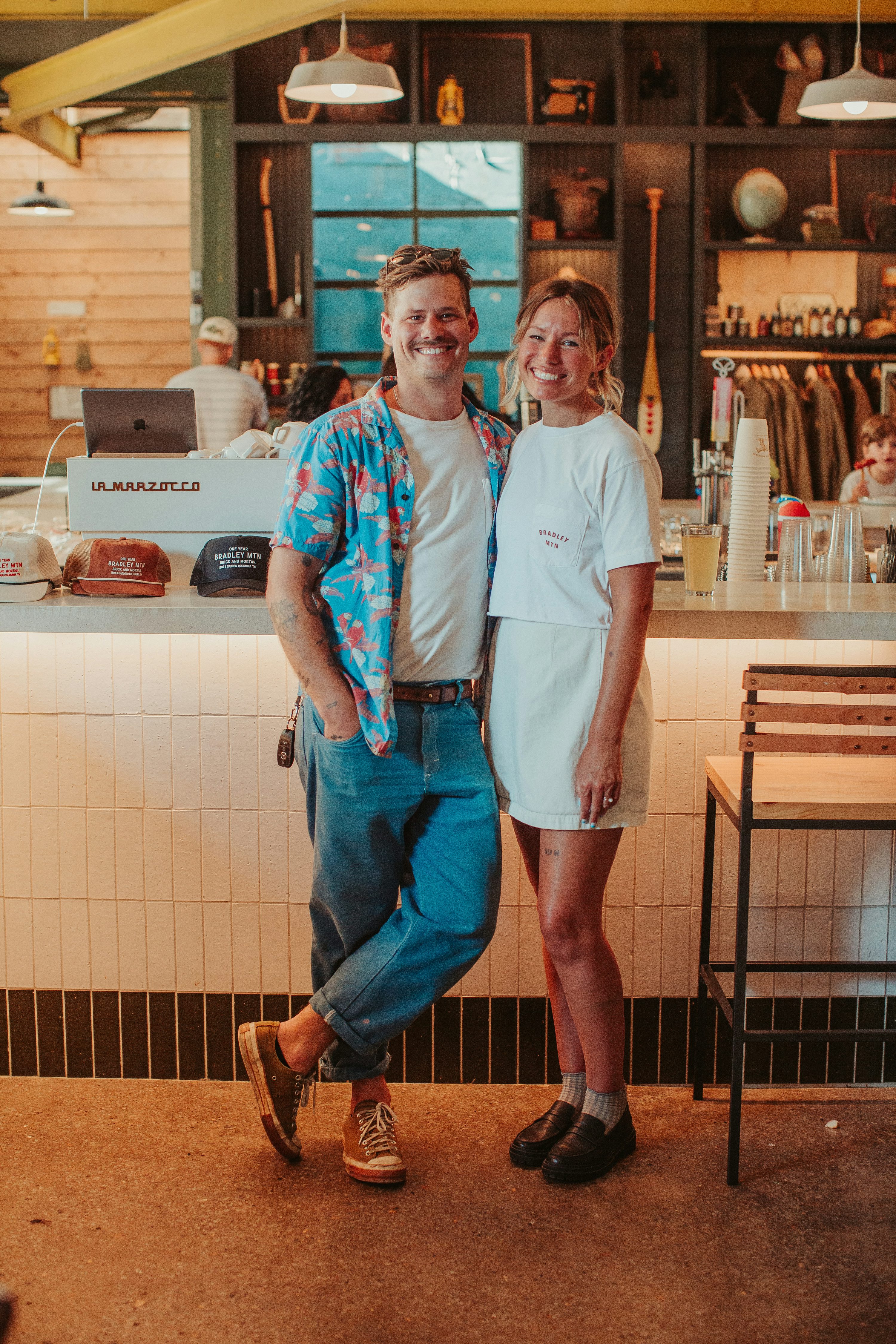 a couple is standing sidebyside in what appears to be a cafe or restaurant setting. the man is wearing a white tshirt, a colorful buttondown shirt, and blue jeans with brown shoes. he is smiling and has a casual, relaxed posture. the woman next to him is wearing a white tshirt and a matching white skirt. she also appears to be smiling. the background shows a counter with bar stools, a coffee machine, and various items displayed on shelves. the lighting is warm and the overall atmosphere seems inviting.