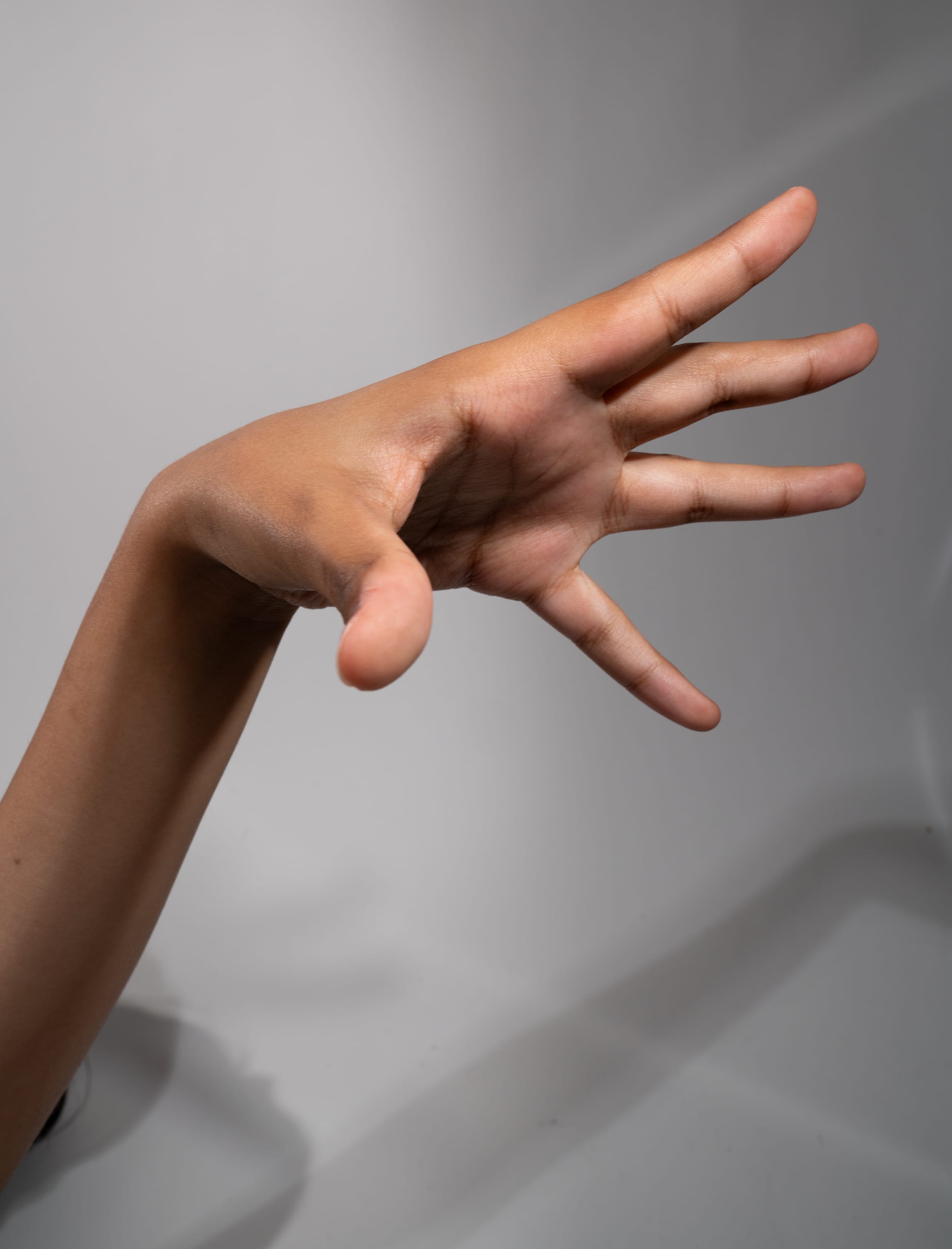 a left hand is extended outwards against a solid white backdrop. the hand is outstretched, with all fingers separated. the arm is slender and has a brown skin tone.