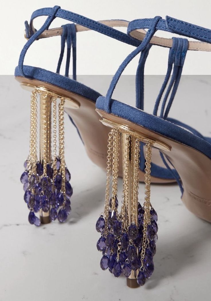 this image features a pair of blue suede sandals with stiletto heels. the heels are adorned with gold chains and purple teardrop beads, adding a decorative element to the shoes. the sandals have an ankle strap and are captured from a back view.