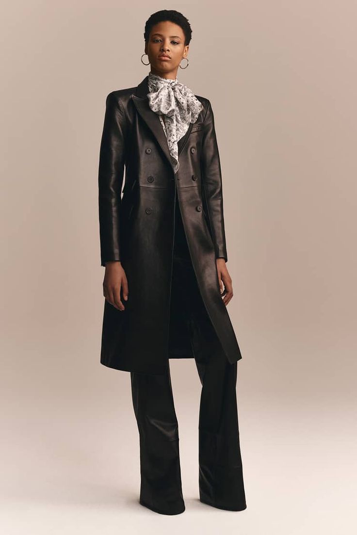a woman model is posing in a studio wearing a long, black leather coat with a double breasted closure. she is wearing a white blouse with a black and white scarf tied around her neck. her wideleg trousers are also black, and she is wearing gold hoop earrings.  she has a serious expression on her face.