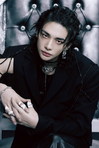 the image is a closeup portrait of hwang hyunjin, a member of the south korean kpop group stray kids. he is styled in a dark, edgy aesthetic.  he's wearing a black blazer with unique detailing, several silver necklaces including a choker, and silver earrings. his long, dark hair is styled messily. his fingers are adorned with small white bandages. he has an intense gaze directed at the camera. the background is a dark, textured, upholstered chair, adding to the overall moody atmosphere of the photograph. the overall style suggests a professional photoshoot with a focus on fashion and a dark, dramatic aesthetic.