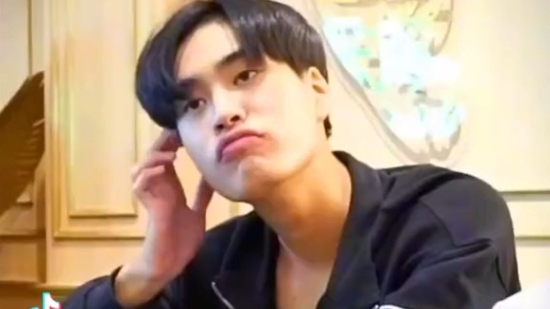 a young asian man with black hair is taking a selfie. he is wearing a black shirt and has a bored expression on his face. he is indoors and the lighting is bright.  the walls behind him are tan and gold with a mural on it.