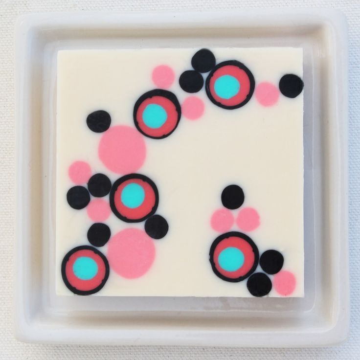 this image shows a bar of handmade soap with a geometric design. the soap is white with pink, black, turquoise, and red circles and dots arranged in a pattern. the soap is sitting in a white soap dish.