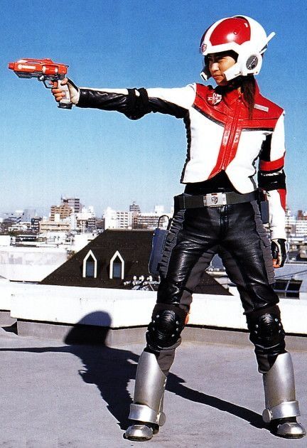 a female superhero in a red, white, and black suit stands on a rooftop with a gun in her hand. she is wearing a futuristic helmet and has a determined look on her face.  the backdrop is an urban cityscape.