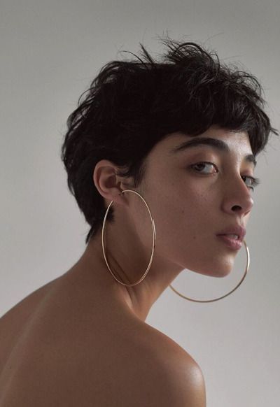 a closeup portrait of a woman with short, dark, tousled hair, showcasing a pixie cut. she is wearing large gold hoop earrings and her bare shoulders are visible. the background is a neutral beige, creating a minimalist aesthetic. the lighting is soft and natural, highlighting her androgynous features and natural beauty.