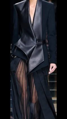 a woman in a black leather jacket with a draped collar and an asymmetric design. the jacket is paired with a black dress that is sheer underneath. the woman is on a runway and looks very sophisticated and chic.