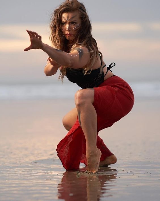 a woman in a red dress stands on a sandy beach with the ocean in the background. she is wearing a black top and has long brown hair. the woman is looking directly at the camera.