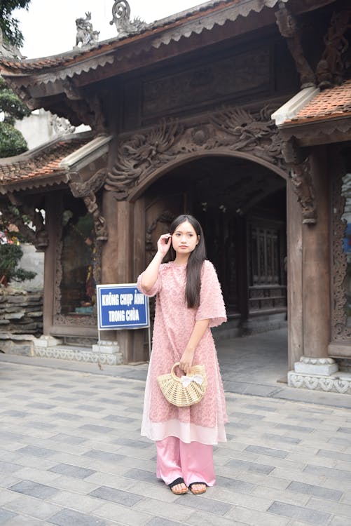 a young woman is standing in front of a temple entrance. she is wearing a pink ao dai, a traditional vietnamese outfit, and carrying a small woven basket. behind her is a wooden temple entrance with ornate carvings. a sign with blue lettering is visible to the left of the woman. the ground is paved with grey stones.