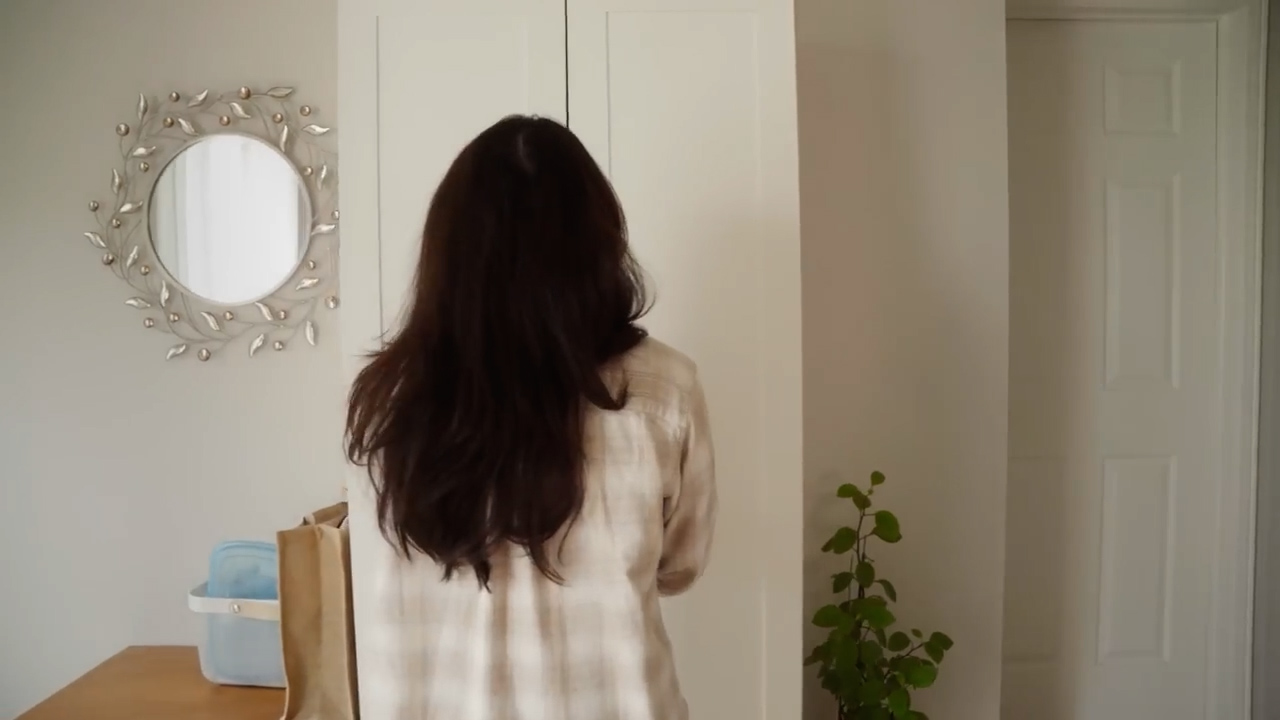 a woman with long, dark brown hair stands with her back to the camera. she is in a room with white walls and a wooden table. there is a round mirror with a silver frame on the wall to the left, and a white door on the right. a small plant sits on the floor in front of the door. the woman is holding a beige bag in her right hand. there is a blue container on the table behind her. the overall atmosphere of the image is calm and serene.