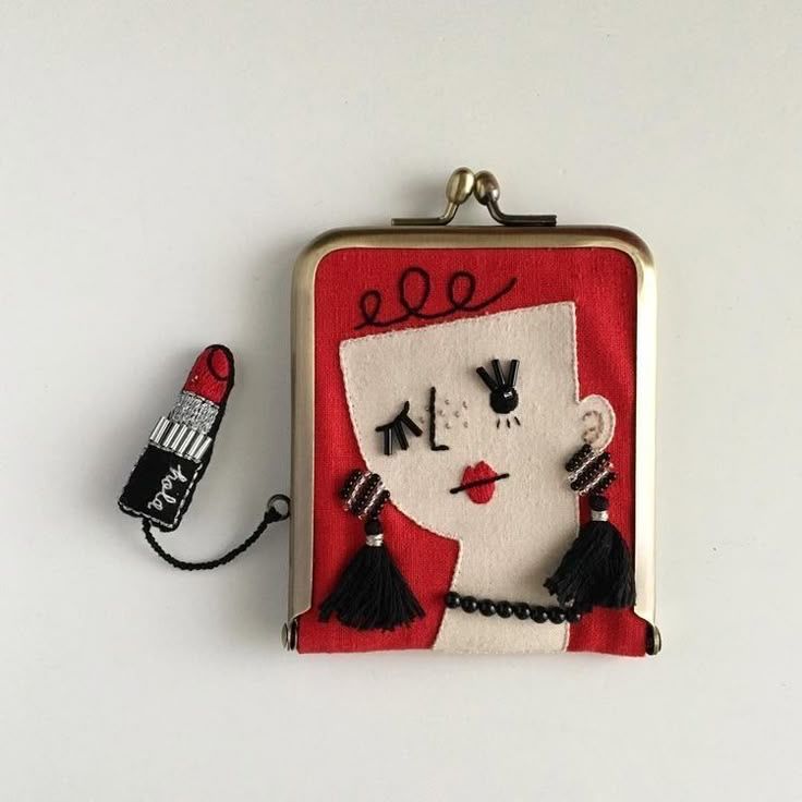 a closeup shot of a rectangular coin purse with a metal frame. the purse is embroidered with a stylized woman's face on a red background. the face features black beaded eyelashes, eyebrows, lips, and a necklace. she wears beaded earrings with black tassels. a small lipstick charm is attached to the side of the purse with a cord.