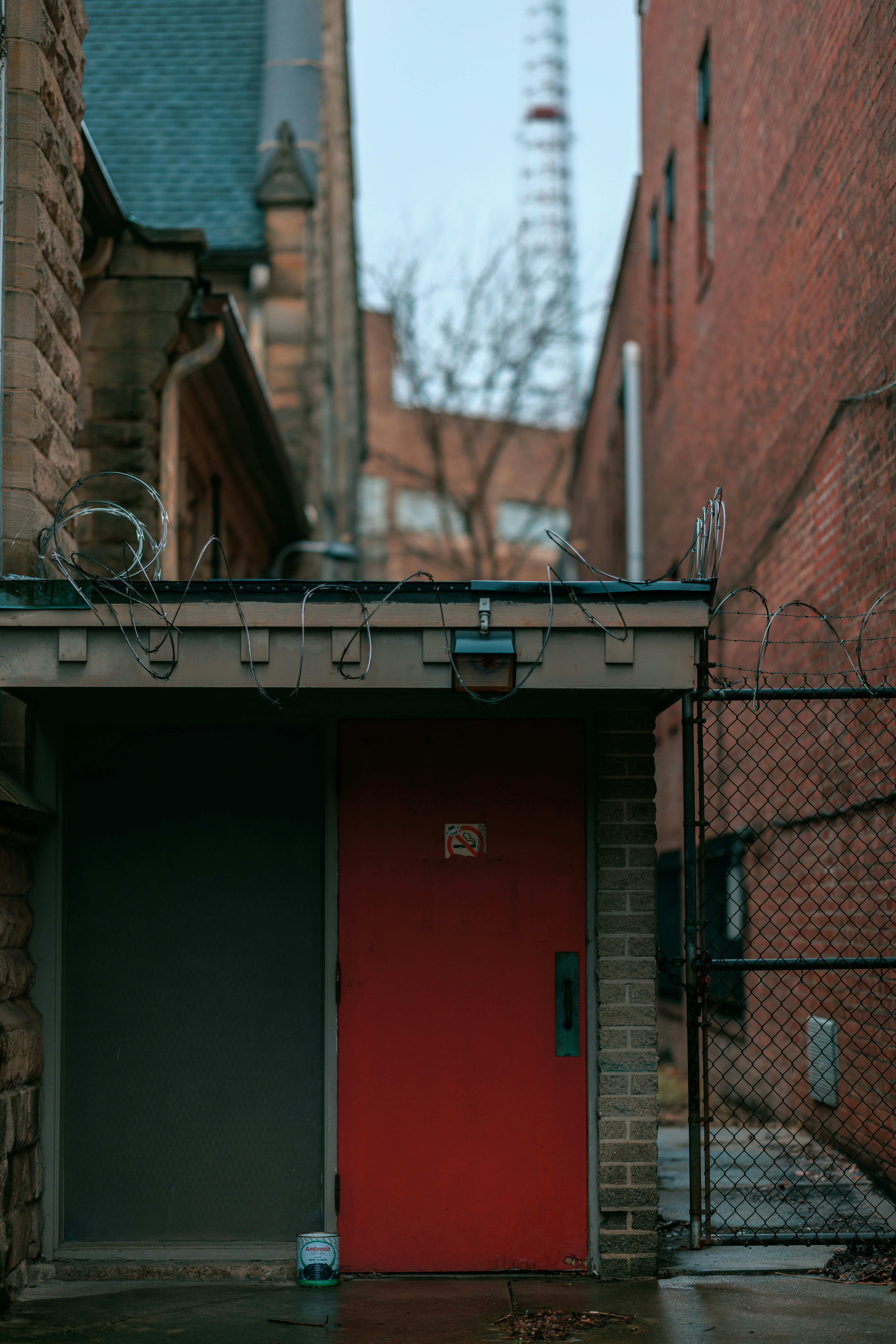 an alleyway between two brick buildings on an overcast day. the focus is drawn to a red door framed by a chainlink fence and topped with barbed wire. the scene exudes a gritty, urban atmosphere.