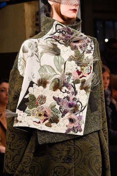 loseup shot of a woman's model showcasing a richly detailed coat. the coat features an intricately embroidered offwhite panel depicting a floral design with feathers and lace accents. it is layered over a dark green paisley patterned coat. the model is wearing a black veil. the background is blurred, indicating a fashion runway setting. the overall aesthetic is highfashion and elegant.