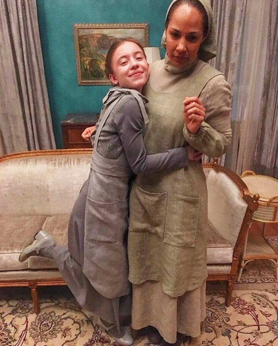 the image shows two actresses, one adult and one child, embracing on a movie or television show set.  they are wearing period costumes reminiscent of the 1920s or 1930s, with simple, mutedcolored dresses and aprons. the child actress is hugging the adult actress from the side, her leg casually draped over the ornate sofa in the background. the set features an antiquestyle living room, complete with a plush light beige sofa and a wicker side table. the overall ambiance suggests a period drama or historical film production.  the background is a tealcolored wall, and a framed landscape painting is visible.