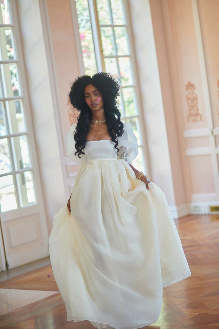 a young woman with dark curly hair is wearing a beautiful, flowing white empire waist gown with puff sleeves.  she is posed in a grand, lightfilled interior space, possibly a palace or mansion, with large windows and hardwood floors. the dress is elegant and romantic. the woman is wearing gold jewelry. the overall style suggests a bridal or wedding theme.