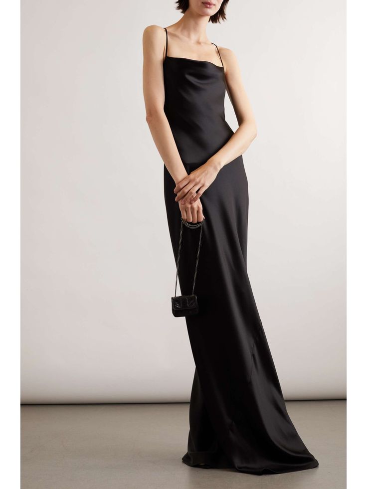 a woman in a black satin dress with a cowl neck poses in a studio with a white background. the dress is long and slimfitting. the woman is wearing high heels and a small clutch bag.