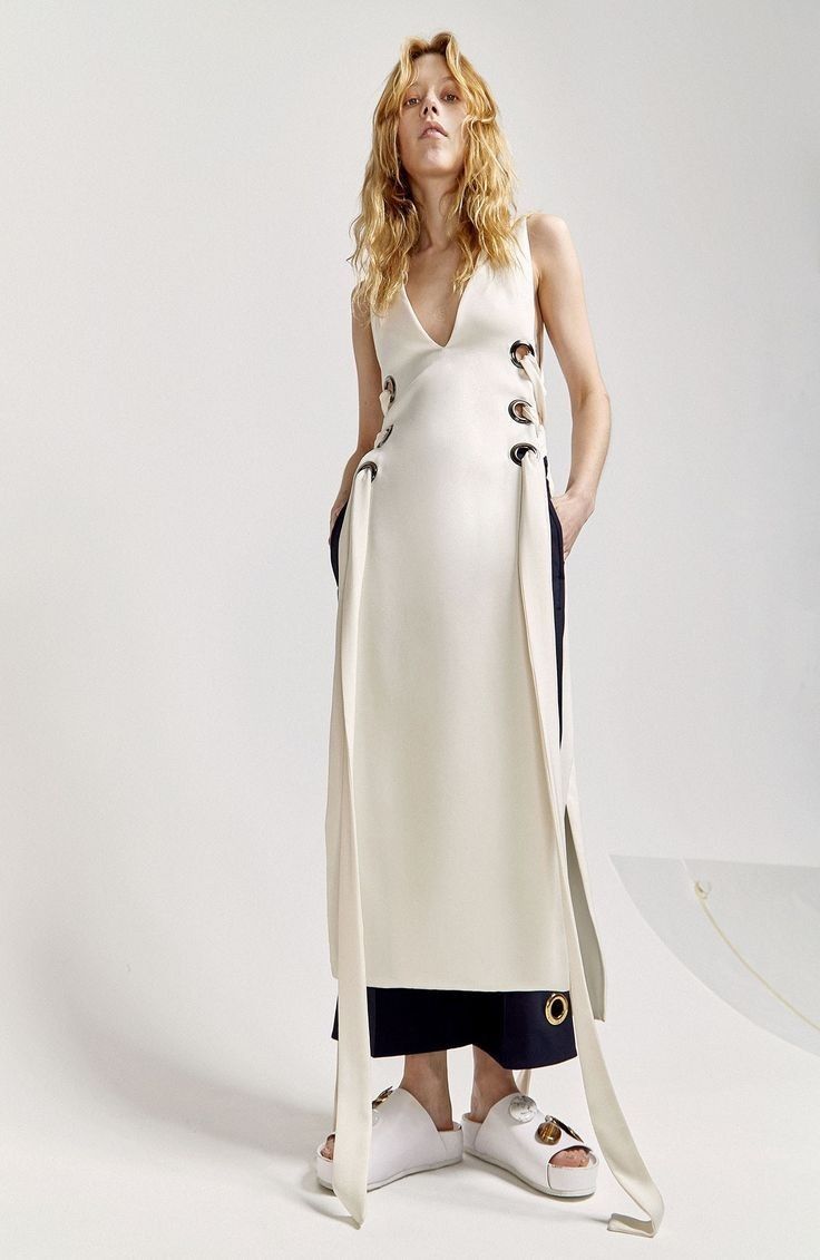a full shot of a tall, slender woman posing in a studio. she has blonde curly hair, fair skin, and is wearing a sleeveless, creamcolored dress with a deep vneck. large grommets with fabric loops are attached to the sides of the dress. underneath the dress, she is wearing dark blue pants. she is wearing thicksoled, white sandals. the background is a bright white studio setting.