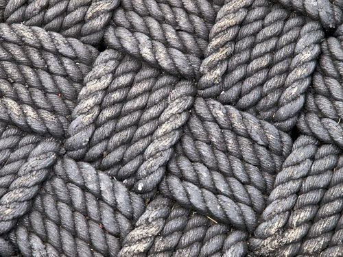 this is a closeup image of a woven rope. the rope is dark grey and black in colour, and it is woven in a tight, intricate pattern. the texture of the rope is rough and coarse, and the individual strands are clearly visible.