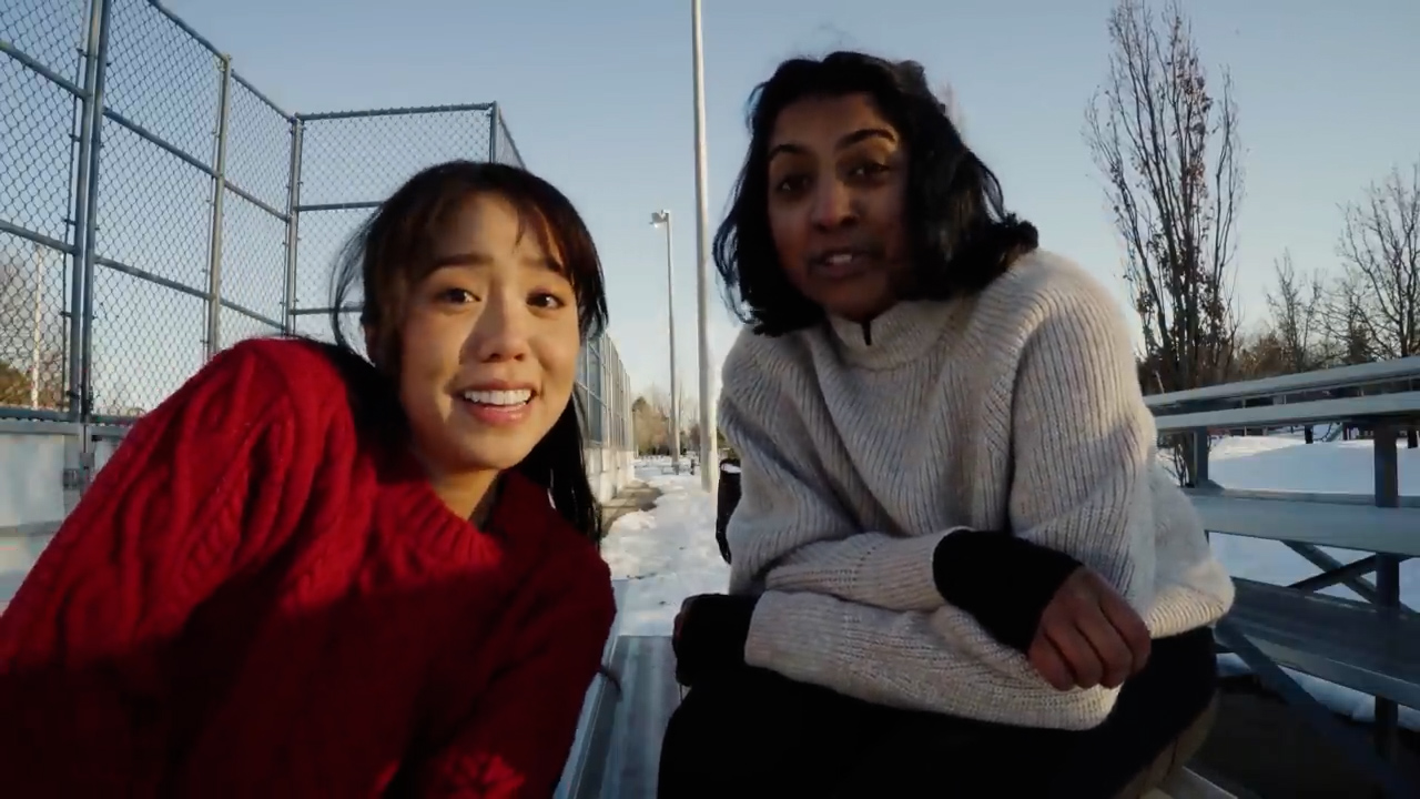 two young women wearing winter clothes are taking a selfie on bleachers in a snowy park. they are both smiling and looking at the camera. the sun is shining, casting shadows on the ground. the overall tone of the image is happy and carefree.