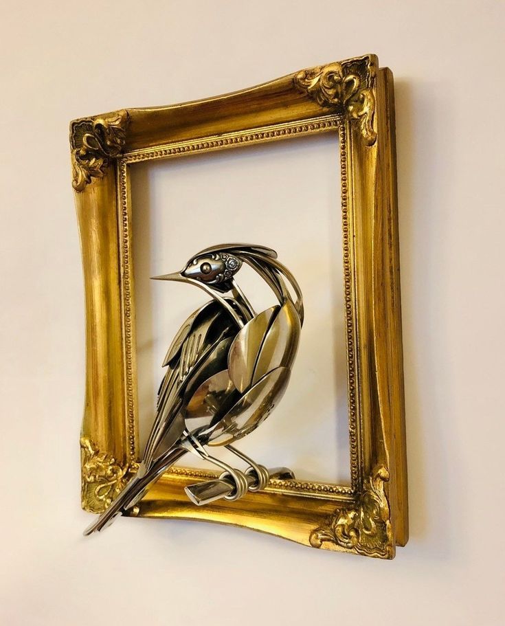 this is a sculpture of a bird made from silverware, which is framed in a gold frame. the bird is perched on a branch. the sculpture is very detailed and intricate, and it is a beautiful example of metalwork.