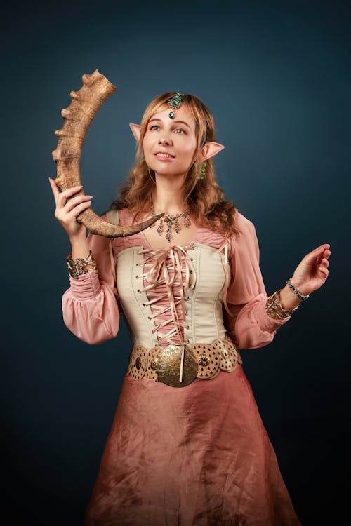 a woman in an elf costume is standing against a dark blue background, holding a large horn. she has fair skin and long, wavy hair. her elf ears are pointed and protrude from her hair. she's wearing a pink dress with a white bodice laced up in the front. she also has a decorative belt with a large gold buckle. her jewelry includes a green gemstone on her forehead, a necklace, bracelets, and earrings. the horn she is holding is large and curved. she is looking up and to the left with a slight smile.