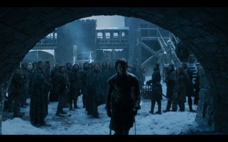 a low angle shot of jon snow (kit harington) standing in the midst of a group of soldiers in the snowy courtyard of winterfell. the scene is dark and cold, emphasizing the harsh winter conditions. jon is framed by a stone archway, creating a sense of claustrophobia and isolation. the image evokes the atmosphere of the popular hbo television series game of thrones.