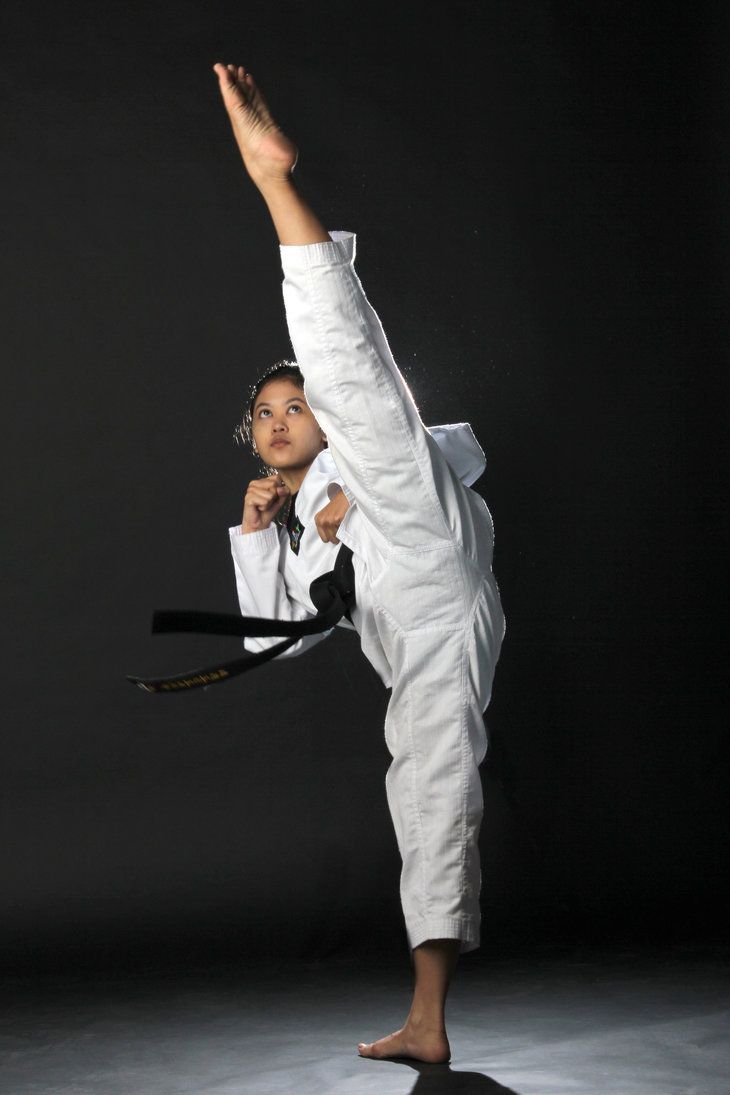 a young woman in a white martial arts uniform and black belt performs a high kick in a darkened studio. she has her leg raised high and is looking straight ahead, her expression focused and determined. she is demonstrating the power and strength of martial arts.