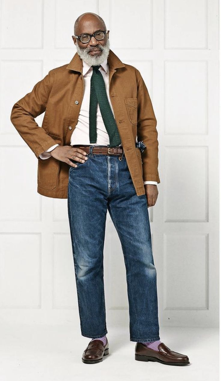 a full shot of an older african american man with glasses and a gray beard, wearing a brown jacket, light pink shirt, green tie, blue jeans, and brown loafers with purple socks. he is standing against a white background with wood paneling.