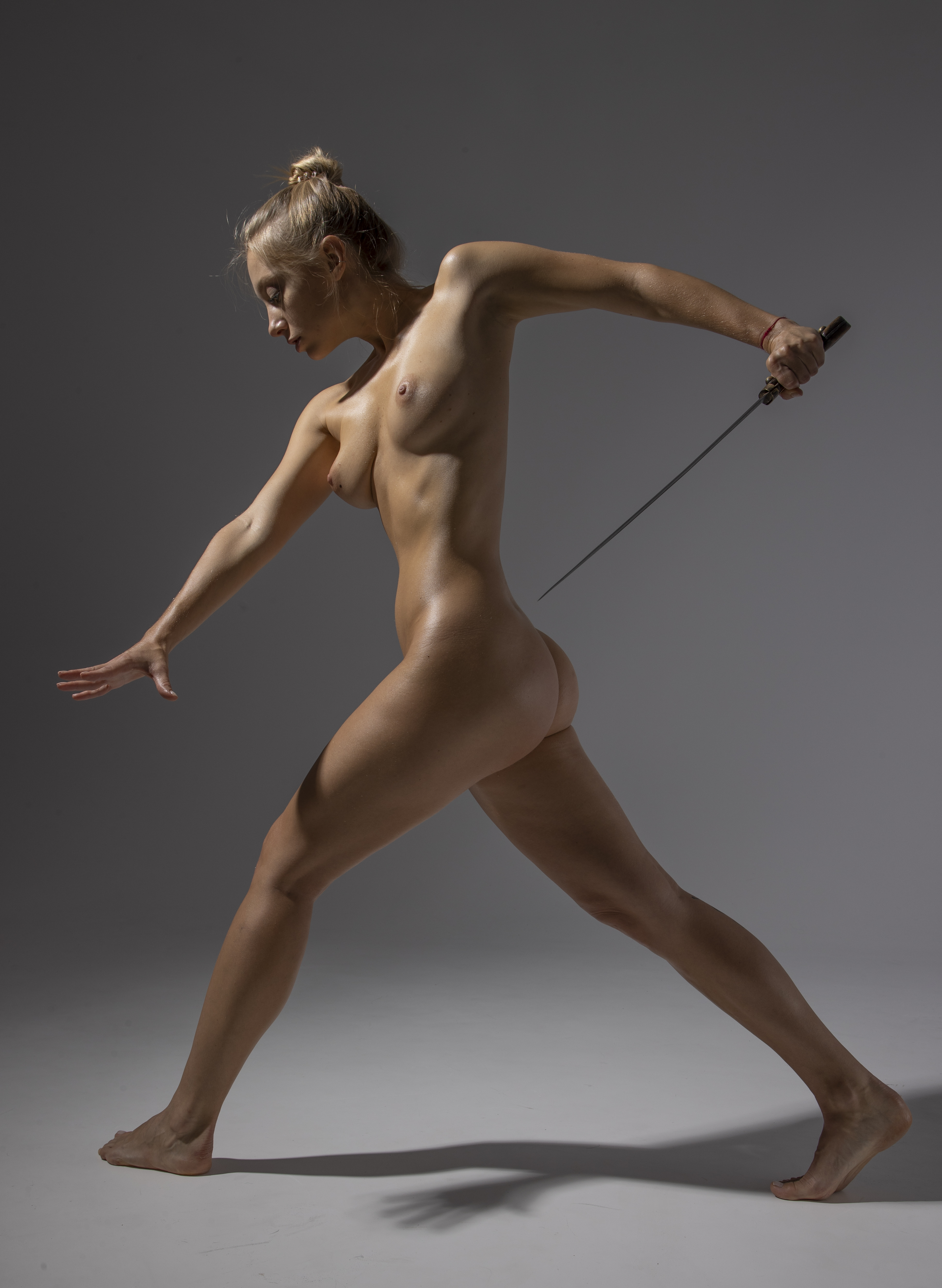 a nude woman is in a warrior pose holding a sword in a studio shot.